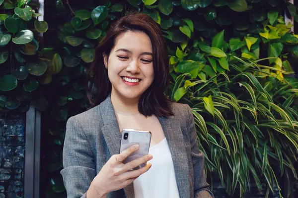 Mutlu, genç, gülümseyen, şık kıyafetler içinde güzel bir Asyalı kadın. Telefondaki mesajları kontrol ediyor. Gri ceketli ve kot pantolonlu.