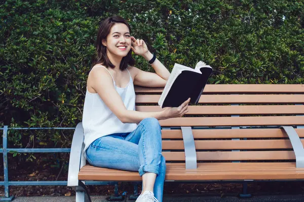 Kahverengi saçlı, kısa boylu, gülen Asyalı bir kadın. Ağaçlara ve çitlere yaslanmış bir kitapla ahşap bankta oturmuş kameraya bakarken gülümsüyor.