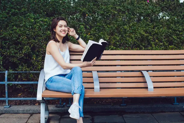 Kahverengi saçlı, kısa boylu, gülen Asyalı bir kadın. Ağaçlara ve çitlere yaslanmış bir kitapla ahşap bankta oturmuş kameraya bakarken gülümsüyor.