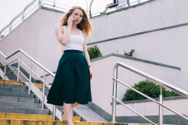 Uzun saçlı, uzun kollu, beyaz gömlekli, yeşil etekli, cep telefonundan arama yapan yaz aylarında merdivenlerden inen sarışın bir kadın.