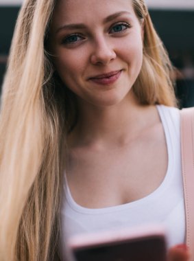 Düşük açılı, beyaz kolsuz tişörtlü, mutlu, genç bir kadın şehirde cep telefonuyla kameraya bakıyor.