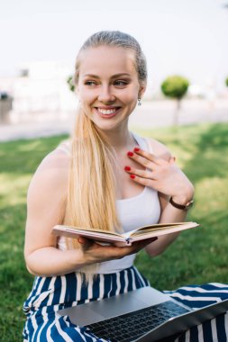 Beyaz kolsuz tişörtlü, çizgili pantolonlu, elinde günlük, dizlerinde dizlerinde dizleriyle yeşil çimlerde oturan genç bir kadın.