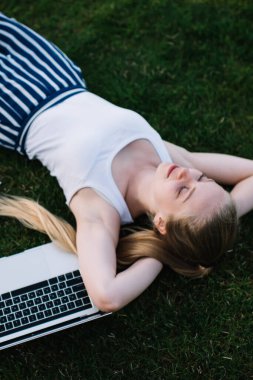 Beyaz kolsuz tişörtlü, çizgili pantolonlu, yeşil çimlerde uzanan ve dizüstü bilgisayarla çalıştıktan sonra rahatlayan genç bayandan.