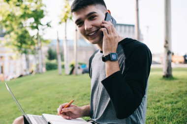 Bilgi kitapçığı ve modern dizüstü bilgisayarı olan neşeli erkek öğrencilerin portresi açık havada oturup danışmanlık için uluslararası sohbetler yapmak ve ders çalışma planı yaratmak için.