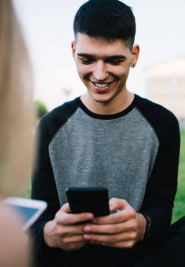 Neşeli gülümseyen içerik, akıllı telefondan mesajlaşan ve arkadaşlarıyla yeşil çimlerin üzerinde rahatlarken sosyal medya kullanan erkek.