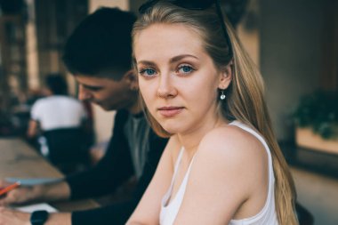 Beyaz kolsuz tişörtlü ciddi bir kadın masada boş siyah ekranlı tabletle oturuyor ve kafe terasında erkek meslektaşıyla buluşurken kameraya bakıyor.