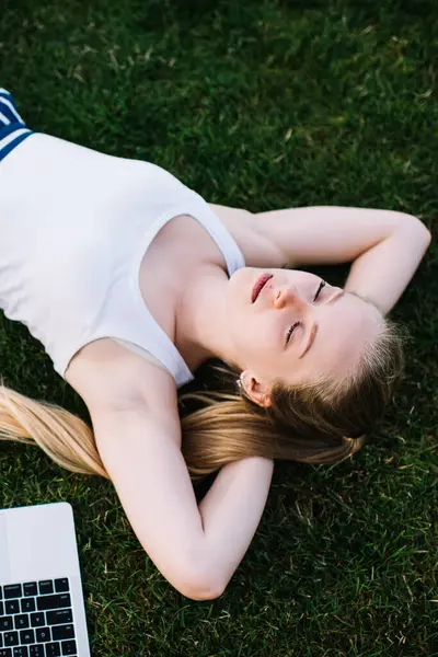 Güzel dijital göçebenin yeşil çimlere uzanmış gözleri kapalı uzanmış seyahat zamanı mesafesinin hayalini kurması çekici kadın serbest çalışanın sakinliği ve dışarıda keyif alması.