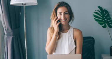 Gündelik giysiler içinde neşeli, modern bir kadın dizüstü bilgisayarı olan bir sandalyede oturmuş kameraya bakıyor ve akıllı telefonuyla konuşuyor.