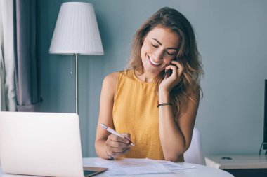 Sıradan giysiler içinde gülümseyen genç bir bayan. Evrakları ve laptopuyla masada oturmuş telefonda konuşuyor.
