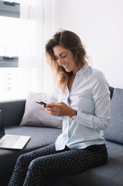 Gömlekli ve pantolonlu yetişkin bir kadın telefonda sohbet ediyor. Oturma odasındaki bilgisayarın yanında oturmuş gülümsüyor.