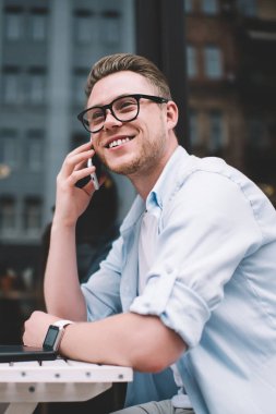 Sıradan gömlekli ve gözlüklü yakışıklı bir genç sokakta masada otururken cep telefonuyla konuşuyor.