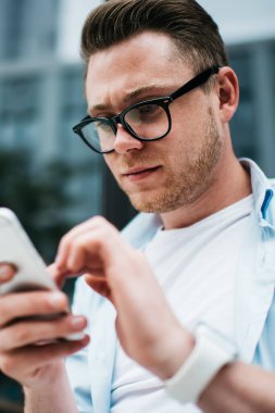 Siyah gözlüklü ve tişörtlü yakışıklı beyaz adamın sokak terasında otururken akıllı telefonuyla sörf yapmasından.