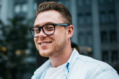 Mavi gömlekli, gözlüklü, cep telefonlu ve caddede otururken kameraya gülümseyen yakışıklı beyaz adam.