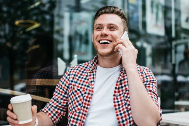 Tişört giyen, beyaz tişört giyen, kahve içen, cep telefonuyla konuşan açık terastaki kalem defteriyle masada oturan mutlu bir erkek.