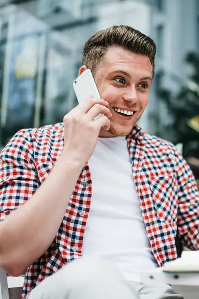 Genç, hayretler içinde, gömleği beyaz, kahve fincanı içen, cep telefonuyla konuşan açık terasta oturan bir erkek.