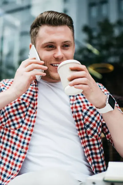 Gündelik giysiler içinde, akıllı saatli mutlu genç bir adam bir fincan kahve içiyor arkadaşlarıyla iletişim kuruyor kafenin dışında telefon dinlendirici kullanıyor.