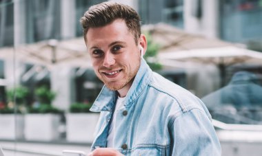 Kot ceketli genç beyaz adam dizüstü bilgisayarı ve cep telefonuyla oturmuş arka planda kameraya gülümsüyor.