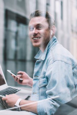 Kot ceketli neşeli milenyum adamı dizüstü bilgisayarı ve cep telefonu ile modern caddede cam çitlerin arkasında oturmuş gülümsüyor.