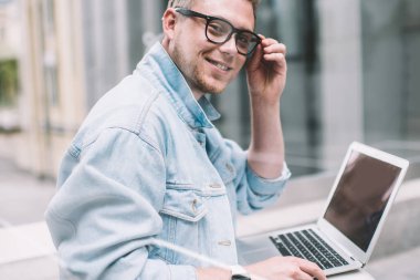 Gözlüklü, kot gömlekli, dizüstü bilgisayarlı ve cam çitin arkasındaki kameraya gülümseyen yakışıklı modern adamın yan görüntüsü.