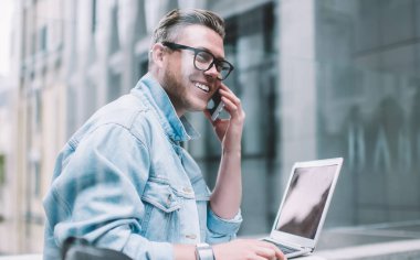 Gündelik giyinmiş, dizüstü bilgisayarlı, akıllı telefon kullanan ve şehir sokaklarında otururken iş arkadaşlarıyla konuşan gülümseyen genç bir adamın camından bakın.