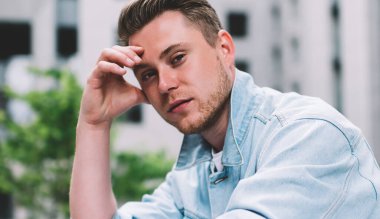 Modern kot ceket giyen, camdan çitlere yaslanan ve sokaktaki kameraya bakan binlerce yıllık beyaz adamın yan görüntüsü.