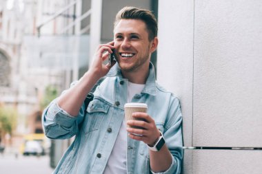 Gündelik giysiler içindeki genç adam cep telefonuyla konuşuyor ve kahve içerken gülüyor ve sokakta dikiliyor.