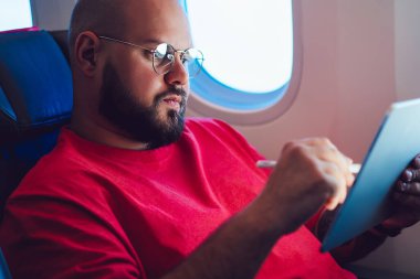 Beyaz erkek uçak yolcusu, kabinde uçak penceresinin yanında otururken tablet ekranına dijital kalemle dokunarak uçağın kablosuz internet bağlantısıyla uçuşunun keyfini çıkarıyor.