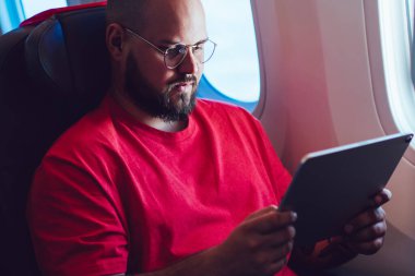 Beyaz erkek uçak yolcusu, elinde tabletle birinci sınıf kabinde uçak penceresinin yanında oturuyor, erkek, tablet kullanırken kablosuz internet bağlantısı ile uçuşunun keyfini çıkarıyor.
