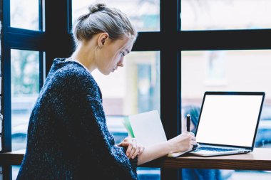 Pencere yakın tek başına oturma modern kahve dükkanı iç masada okurken genç sarışın saç kadın yan görünüm sahte ekran ile dizüstü bilgisayar kullanarak. Freelance üzerinde çalışan genç çağdaş kadın