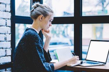 Pencere yakın tek başına oturma modern kahve dükkanı iç masada okurken genç sarışın saç kadın yan görünüm sahte ekran ile dizüstü bilgisayar kullanarak. Freelance üzerinde çalışan genç çağdaş kadın