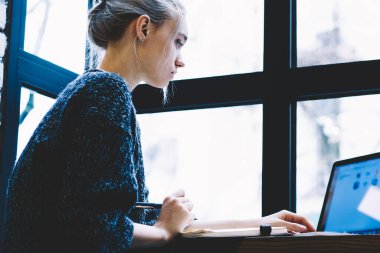 Pencere yakın yalnız oturan modern kahve dükkanı iç masada okurken dizüstü bilgisayar kullanarak Casual giysiler genç sarışın saç dişi yan görünümü. Freelance üzerinde çalışan genç çağdaş kadın