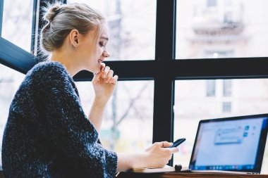 Modern kahve dükkanı iç penceresine karşı masada dizüstü bilgisayar ve çalışmalar ile otururken akıllı telefon kullanarak çağdaş genç sarışın saç kadın Freelancer yan görünümü. Milenyal insanlar ve teknoloji