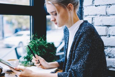 Modern güzel sarışın saç kadın yan görünüm masada kahve dükkanı pencere yakın tablet kullanarak ve Not defteri 'nde not alarak oturuyor. Hipster kız Blogger oluşturma Makale