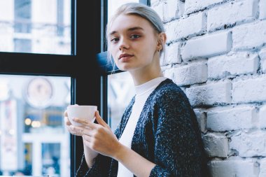 Modern kafe iç kapuçino tutmak ve kamerada güvenle görünümlü masada oturan Casual giysiler Klasik Genç 20 yaşındaki kadın yan görünümü. Kadın Freelancer