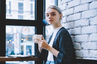 Modern kafe iç kapuçino tutmak ve kamerada güvenle görünümlü masada oturan Casual giysiler Klasik Genç 20 yaşındaki kadın yan görünümü. Kadın Freelancer