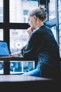 Rahat dalgın genç kadının yan görünümü pencere ve sörf laptop karşı masada modern kafeterya oturan kahve fincan varken. Düşünceli kadın Blogger bilgisayar üzerinden Freelance üzerinde çalışıyor