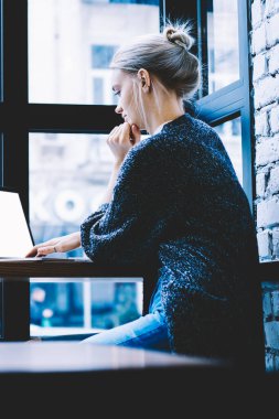 Pencere yakın tek başına oturma modern kahve dükkanı iç masada okurken genç sarışın saç kadın yan görünüm sahte ekran ile dizüstü bilgisayar kullanarak. Freelance üzerinde çalışan genç çağdaş kadın