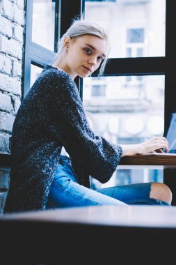Yan görünüm genç Casual sarışın saç hipster kız laptop ve Not defteri ile masada kafede yakın pencerede kamera bakıyor oturuyor. Kahve dükkanı iç bilgisayar ile online öğrenme kadın öğrenci