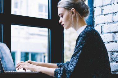 Pencere yakın yalnız oturan modern kahve dükkanı iç masada okurken dizüstü bilgisayar kullanarak Casual giysiler genç sarışın saç dişi yan görünümü. Freelance üzerinde çalışan genç çağdaş kadın