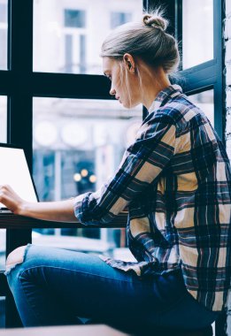 Pencere yakın oturan modern kahve dükkanı iç masada okuyan iken rahat giysiler genç sarışın saç kadın yan görünümü masa başında bilgisayar kullanarak. Freelance üzerinde çalışan genç çağdaş kadın