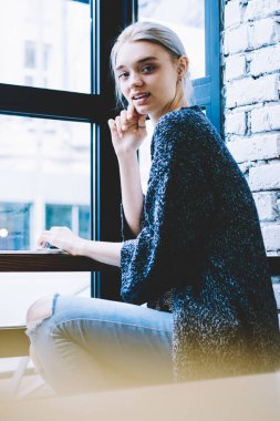 Yan görünüm genç Casual sarışın saç hipster kız laptop ve Not defteri ile masada kafede yakın pencerede kamera bakıyor oturuyor. Kahve dükkanı iç bilgisayar ile online öğrenme kadın öğrenci