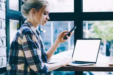 Modern sarışın saç kadın masada oturan cep telefonu uygulama üzerinden Laptop bilgisayar tarama ağı Mock kadar yakın. Millennial hipster kız harcama zaman kullanarak teknoloji içinde kahve dükkanı