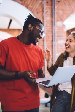 Modern, mutlu, çok kültürlü kadın ve taşınabilir bilgisayarlı bir adam üniversite çalışma odasında yan yana durmuş sohbet ederken mutlu bir şekilde gülüyorlar.