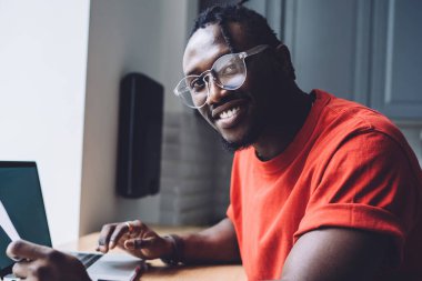 Üzerinde günlük tişört ve gözlüklerle elinde çalışma kağıtları olan gülümseyen Afro-Amerikan 'ın yan görüntüsü ve masada dizüstü bilgisayarla oturan kameraya bakışı.