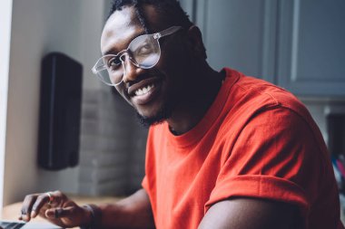 Üzerinde günlük tişört ve gözlüklerle elinde çalışma kağıtları olan gülümseyen Afro-Amerikan 'ın yan görüntüsü ve masada dizüstü bilgisayarla oturan kameraya bakışı.