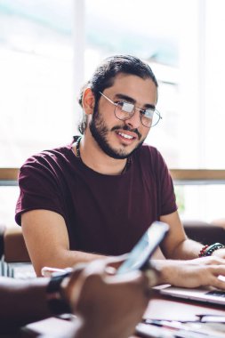 Gözlüklü, gülümseyen, dizüstü bilgisayarda yazan, siyahi bir adamla aynı masada oturan, telefonu kullanan ve kameraya bakan bir adam.