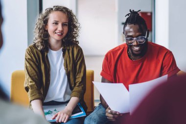 Gülümseyen kıvırcık kadın ve gündelik Afrikalı Amerikalı adam ofis salonunda oturmuş grup arkadaşlarıyla sohbet ediyorlar.