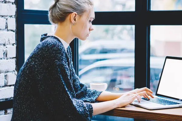 Pencere yakın yalnız oturan modern kahve dükkanı iç masada okurken dizüstü bilgisayar kullanarak Casual giysiler genç sarışın saç dişi yan görünümü. Genç kadın Freelancer