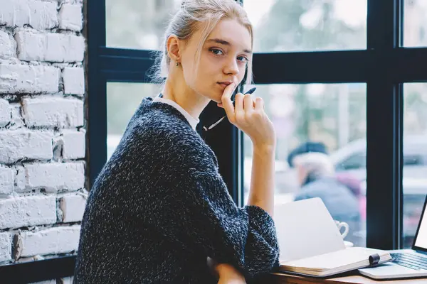 Yan görünüm genç Casual sarışın saç hipster kız laptop ve Not defteri ile masada kafede yakın pencerede kamera bakıyor oturuyor. Kahve dükkanında laptop ile kadın öğrenci öğrenme