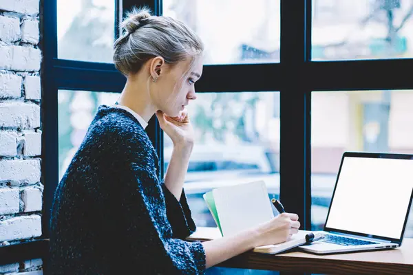 Pencere yakın tek başına oturma modern kahve dükkanı iç masada okurken genç sarışın saç kadın yan görünüm sahte ekran ile dizüstü bilgisayar kullanarak. Freelance üzerinde çalışan genç çağdaş kadın